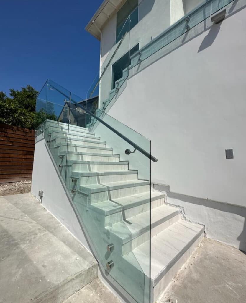 Outdoor staircase featuring a clear glass railing with stainless steel brackets and top rail, set against a modern white exterior
