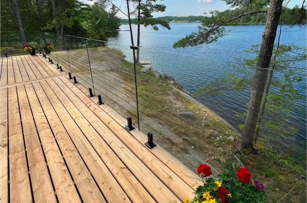 Modern deck yard with installed glass railing beside a lake, framed by trees in bloom