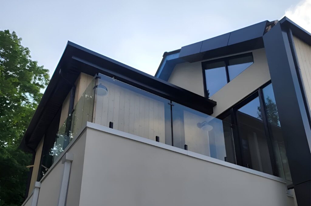Outdoor view of a modern residential house with a mansard design, featuring a newly installed balcony with clear glass railings, showcasing a sleek exterior and contemporary architectural style.