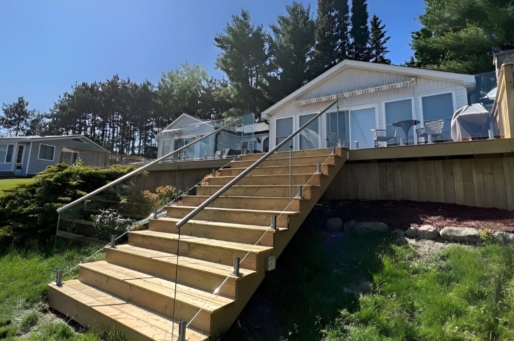 Sunny day view of a modern deck with stairs fully enclosed by high-quality custom glass railings, including glass balustrades along the deck edge, showcasing a clean and durable outdoor design.
