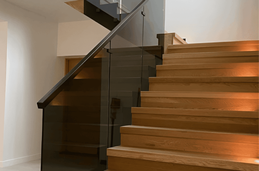 Basement staircase featuring glass railing and warm step lighting for a modern, elegant interior
