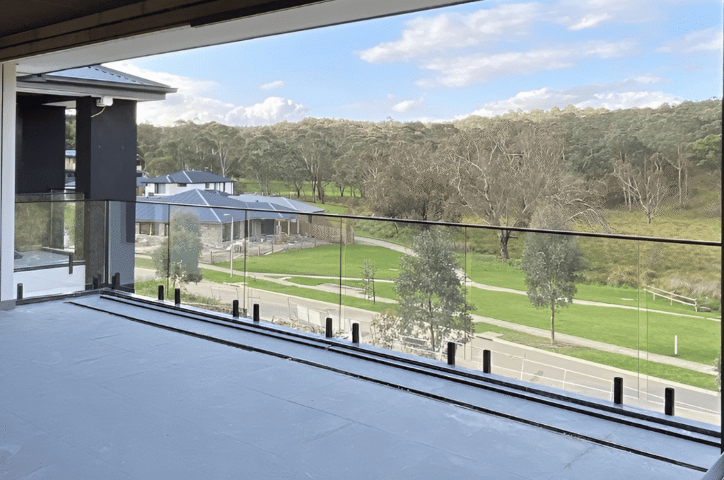 Frameless glass balcony railing with a matte black base shoe channel on a tiled outdoor terrace overlooking a residential neighborhood and green landscape.