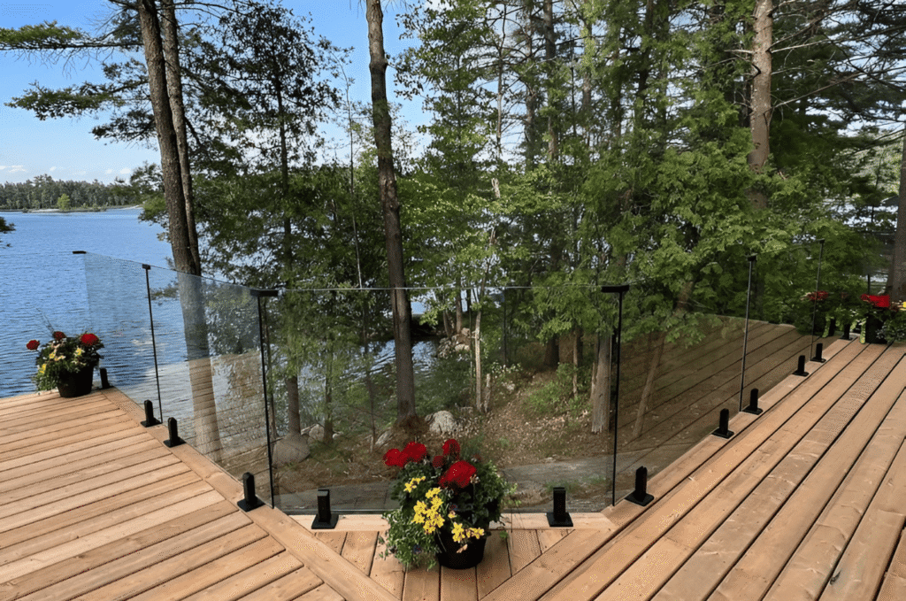 Post-and-clamp glass deck railing with matte black hardware on a waterfront wood deck overlooking a lake and trees.