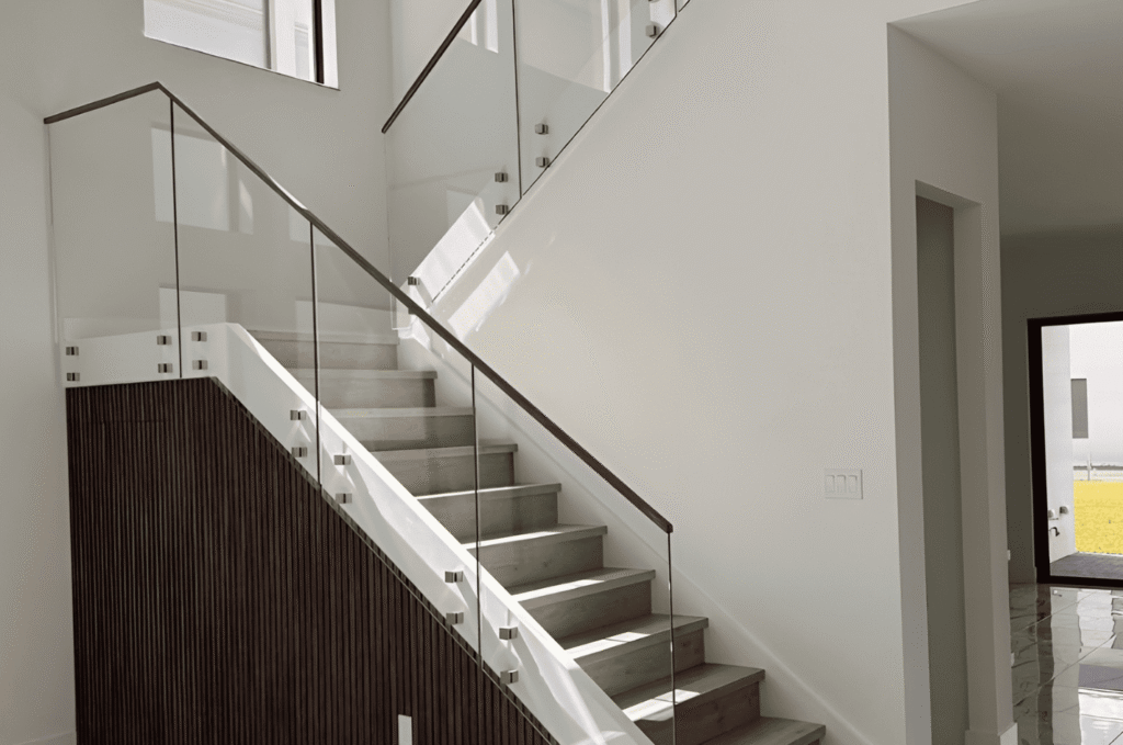 Interior frameless glass railing with standoff hardware alongside stone stair treads in a light-filled modern home. 