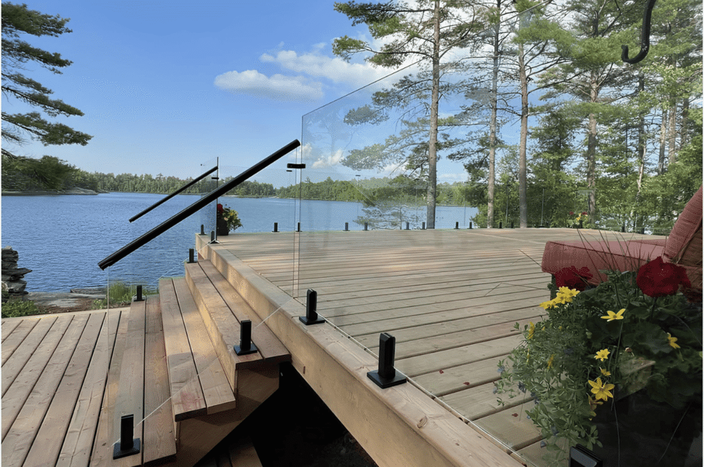 Frameless glass railing installation on a lakeside deck with black standoff hardware, wooden steps, and open water views