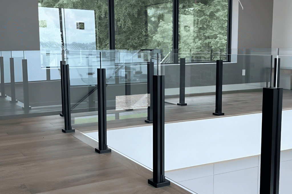 Modern indoor glass railing with black posts beside large windows, connecting open home interiors with bright outdoor views 