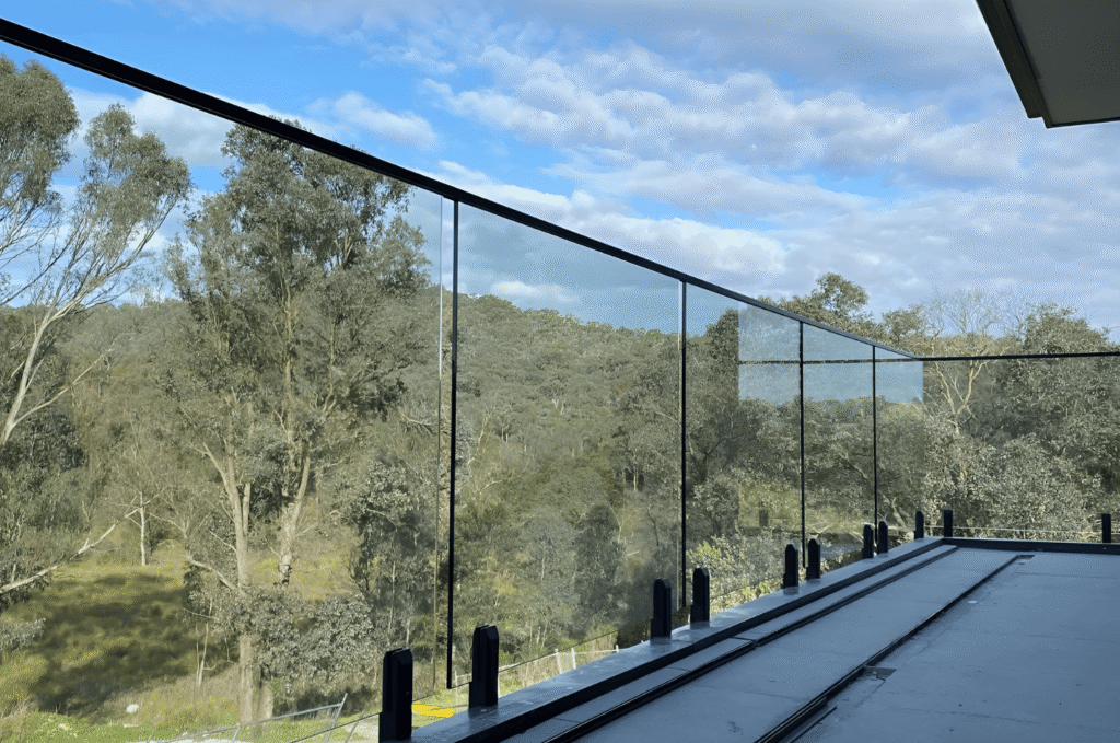 Outdoor glass railing on a modern balcony with clear panels, black posts, and open views across green trees and bright sky 
