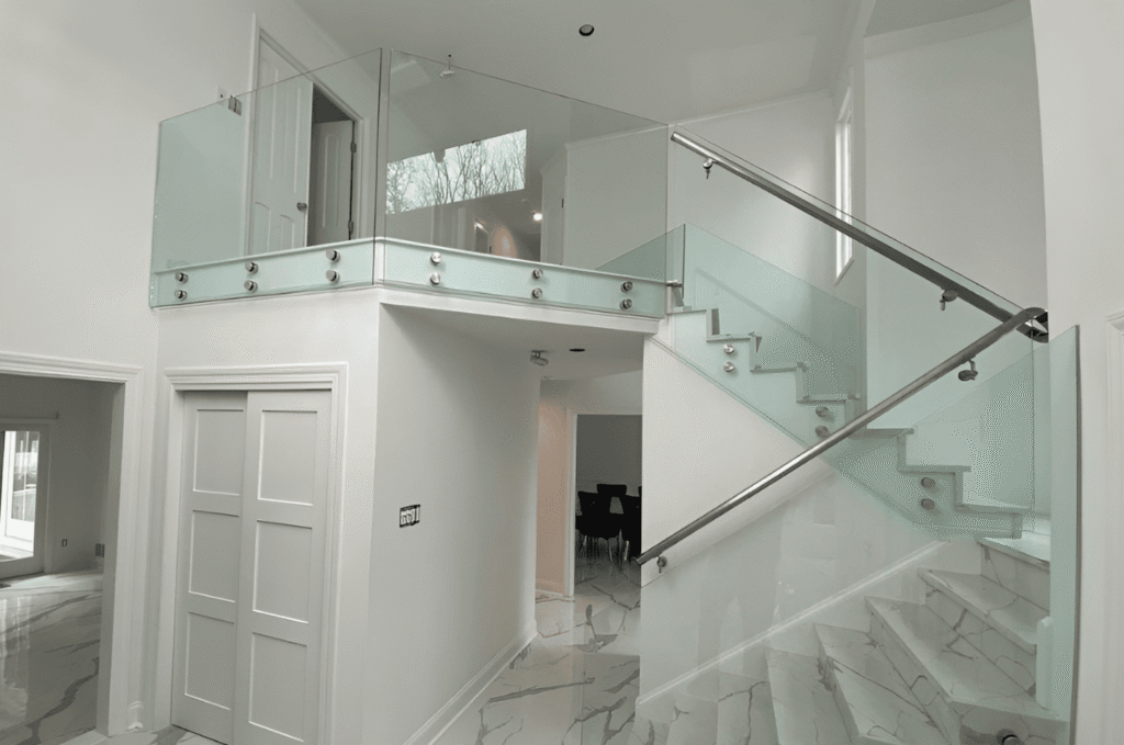 Frameless glass balcony railing with standoffs overlooking marble staircase in modern double height foyer interior design
