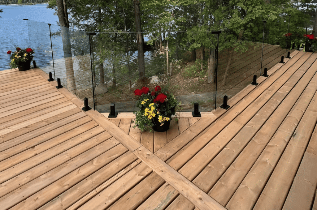 Deck glass railing with black post-and-clamp mounts running along a lakefront wood deck surrounded by trees near Seattle